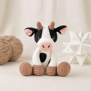 May include: A small, handmade crochet cow plush toy featuring a white body with black spots, a light pink snout, and tan-colored horns and feet. The cow has large, dark, round eyes and pointed ears with pink inner details, sitting in an upright position.