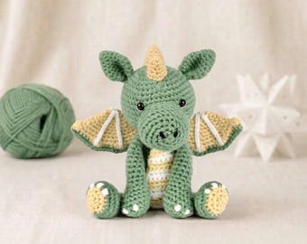 May include: A crocheted dragon plush toy in sage green with pale yellow accents on the wings, belly, feet, and a single horn on its head. The creature features small black safety eyes and a textured yarn construction with visible stitching.