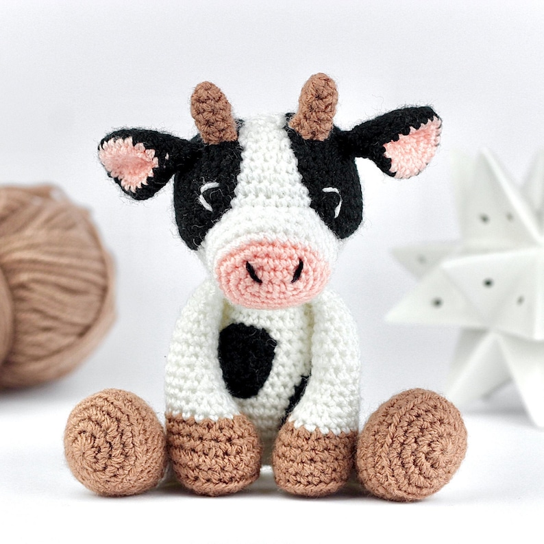 May include: A small, crocheted cow toy featuring a white body with black patches, a light pink snout, and light brown horns and hooves. The cow has black ears with pink inner details and embroidered eyes, sitting in an upright position.