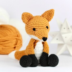 May include: A crocheted fox toy with orange and white fur, black eyes, and black nose. The toy is sitting on a white surface.