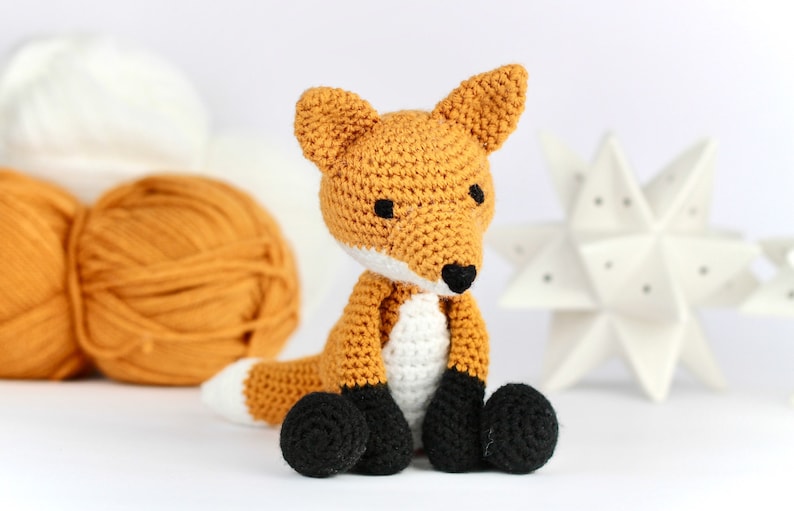 May include: A crocheted fox toy with orange and white fur, black eyes, and black nose. The toy is sitting on a white surface.