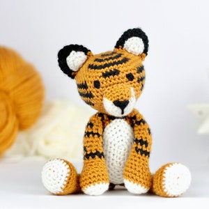 May include: A crocheted tiger stuffed animal with orange and black stripes, white paws, and a white belly. The tiger is sitting on a white surface.
