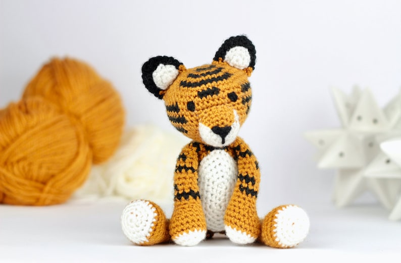 May include: A crocheted tiger stuffed animal with orange and black stripes, white paws, and a white belly. The tiger is sitting on a white surface.