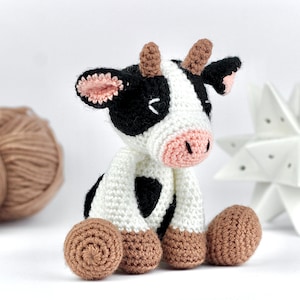 May include: A small, handmade crochet cow toy featuring a white body with black patches, a light pink snout, and brown hooves. The cow has two small brown horns on its head, black and white ears with pink inner details, and closed eyes.