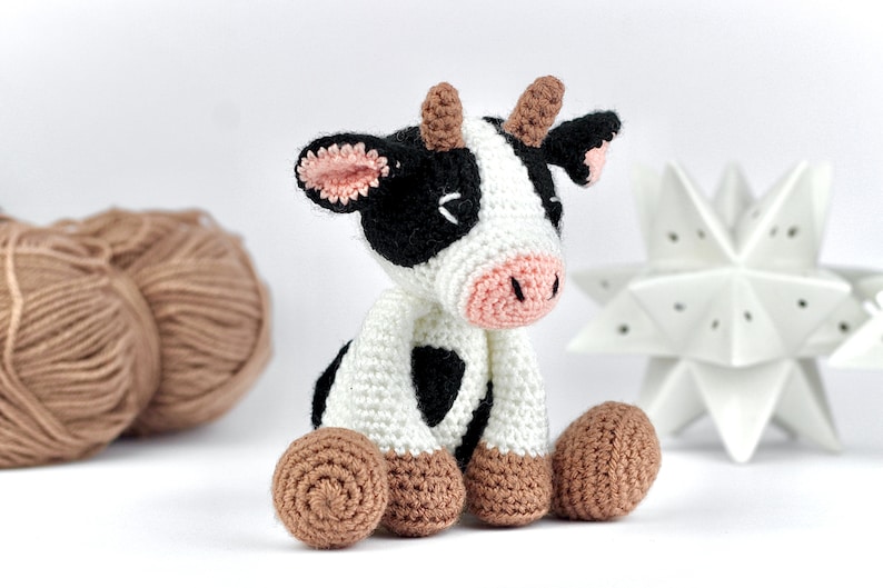 May include: A small, handmade crochet cow toy featuring a white body with black patches, a light pink snout, and brown hooves. The cow has two small brown horns on its head, black and white ears with pink inner details, and closed eyes.