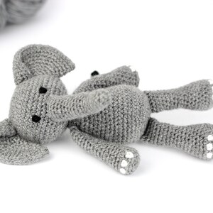 May include: A gray crocheted elephant toy with white accents on its feet. The elephant is lying on its side with its trunk extended.