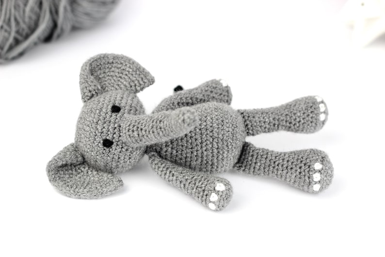 May include: A gray crocheted elephant toy with white accents on its feet. The elephant is lying on its side with its trunk extended.