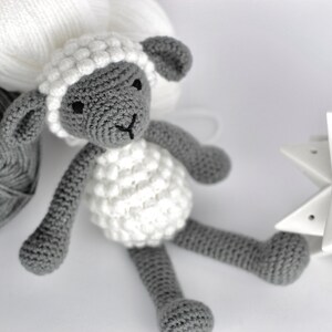 May include: A crocheted gray and white sheep toy with black eyes. The sheep is sitting on a white surface with gray and white yarn balls in the background.
