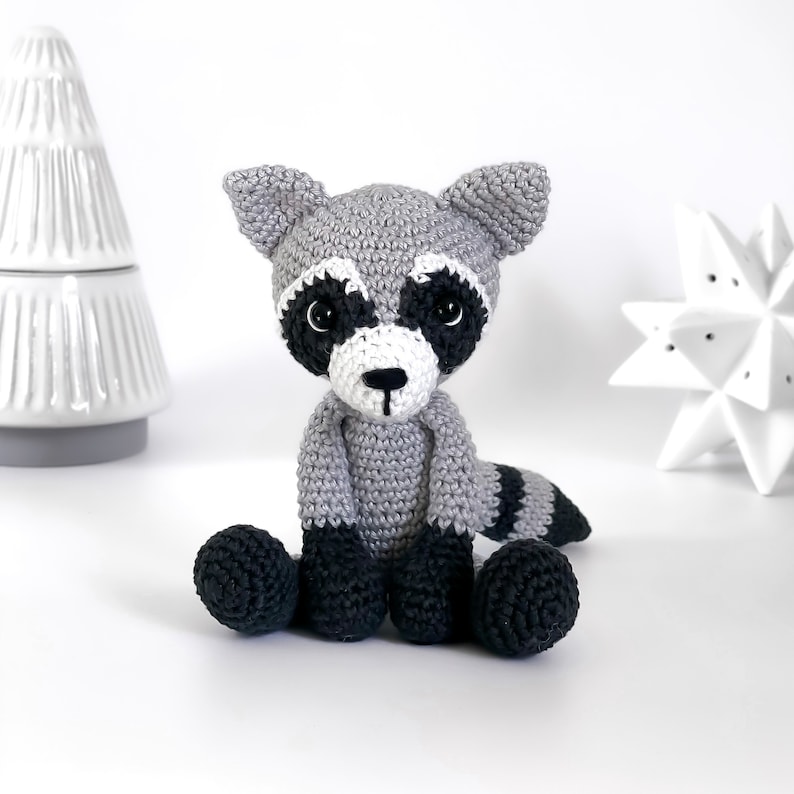 Puede incluir: Un juguete de mapache de crochet con rasgos faciales negros y blancos, sentado sobre una superficie blanca. El juguete es gris con rayas negras en la cola.