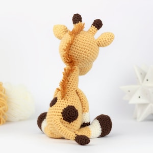 May include: A crocheted giraffe toy with brown spots, sitting on a white surface. The giraffe is made with beige yarn and has brown ears and hooves.