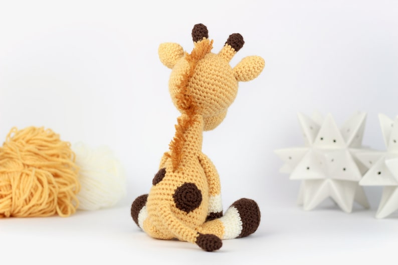 May include: A crocheted giraffe toy with brown spots, sitting on a white surface. The giraffe is made with beige yarn and has brown ears and hooves.