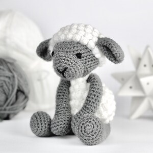 May include: A crocheted gray and white sheep toy with a fluffy white fleece. The sheep is sitting on a white surface.