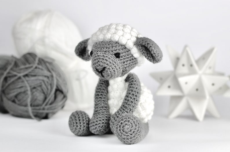 May include: A crocheted gray and white sheep toy with a fluffy white fleece. The sheep is sitting on a white surface.