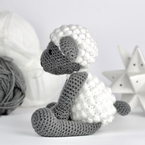 May include: A crocheted gray and white sheep toy with a fluffy white fleece. The sheep is sitting on a white surface.