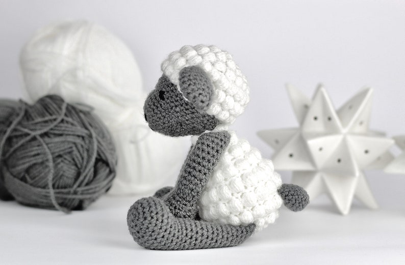May include: A crocheted gray and white sheep toy with a fluffy white fleece. The sheep is sitting on a white surface.