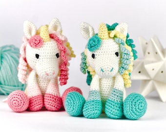 Könnte beinhalten: Zwei gehäkelte Einhorn-Spielzeuge, eines mit rosa und gelbem Garn und das andere mit grünem und gelbem Garn. Beide Einhörner haben weiße Körper und schwarze Augen.