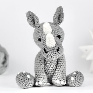 May include: A gray crocheted rhinoceros toy with white accents. The toy is sitting on a white surface.