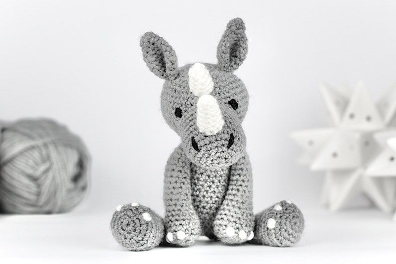 May include: A gray crocheted rhinoceros toy with white accents. The toy is sitting on a white surface.