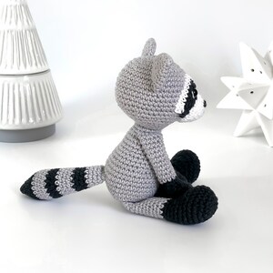 Puede incluir: Un juguete de mapache de crochet sentado en una superficie blanca. El mapache es gris con rayas negras en la cola y orejas y ojos negros.