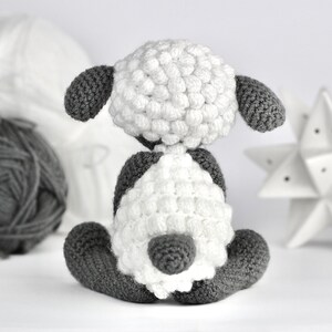 May include: A white crocheted sheep with gray ears and legs. The sheep is sitting on a white surface.