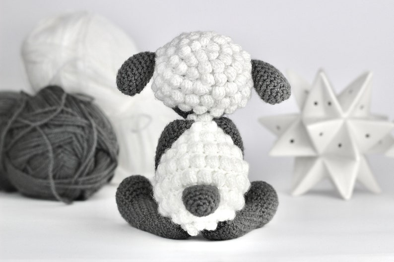 May include: A white crocheted sheep with gray ears and legs. The sheep is sitting on a white surface.