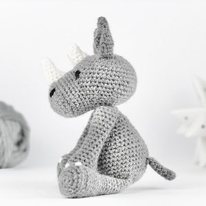 Pode incluir: Um brinquedo de pel&uacute;cia de rinoceronte cinza de croch&ecirc; com chifres brancos e uma cauda branca. O brinquedo est&aacute; sentado em uma superf&iacute;cie branca.