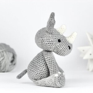 Pode incluir: Um brinquedo de rinoceronte de croch&ecirc; cinza com detalhes brancos nos chifres e p&eacute;s. O brinquedo est&aacute; sentado em uma superf&iacute;cie branca.