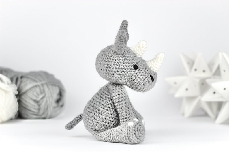 Pode incluir: Um brinquedo de rinoceronte de croch&ecirc; cinza com detalhes brancos nos chifres e p&eacute;s. O brinquedo est&aacute; sentado em uma superf&iacute;cie branca.