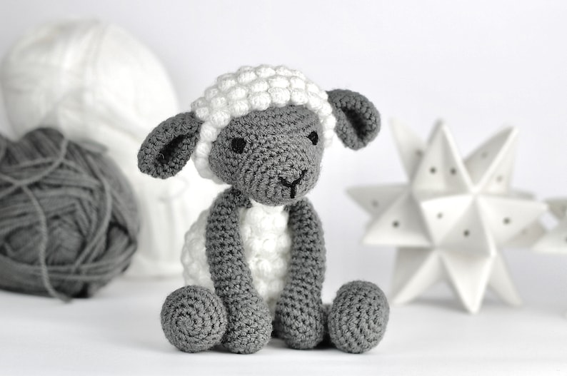 May include: A crocheted gray and white sheep toy with black eyes. The sheep is sitting on a white surface.