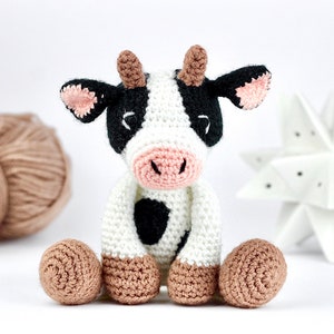 May include: A crocheted stuffed cow toy with black and white spots, brown ears, and a pink nose. The cow is sitting with its legs crossed and has a friendly expression.