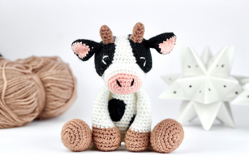 May include: A crocheted stuffed cow toy with black and white spots, brown ears, and a pink nose. The cow is sitting with its legs crossed and has a friendly expression.
