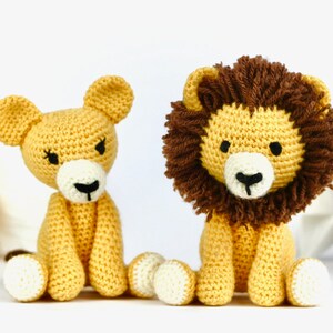 May include: Two crocheted lion plushies, one with a brown mane and one without, sitting on a white surface. Both plushies are yellow with white feet and black eyes.