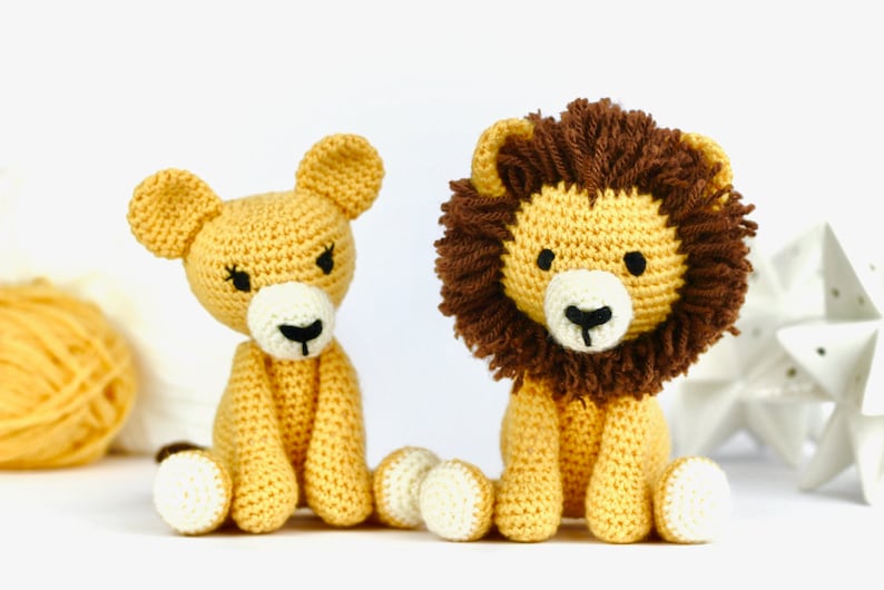 May include: Two crocheted lion plushies, one with a brown mane and one without, sitting on a white surface. Both plushies are yellow with white feet and black eyes.