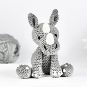 Pode incluir: Um brinquedo de rinoceronte de croch&ecirc; cinza com detalhes brancos sentado em uma superf&iacute;cie branca. O brinquedo tem um chifre branco e marcas brancas no rosto.