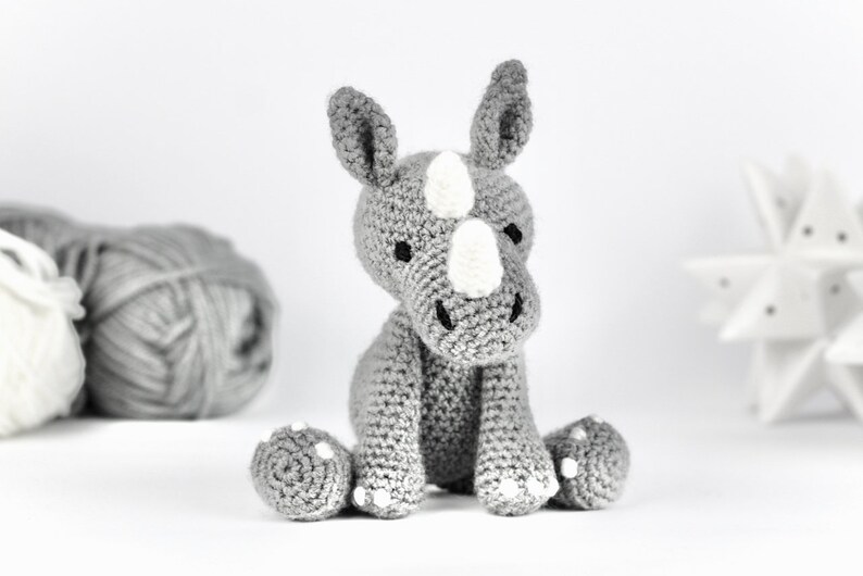 Pode incluir: Um brinquedo de rinoceronte de croch&ecirc; cinza com detalhes brancos sentado em uma superf&iacute;cie branca. O brinquedo tem um chifre branco e marcas brancas no rosto.