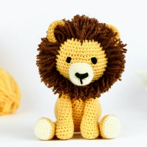 May include: A crocheted lion toy with a brown mane, yellow body, and white face. The lion is sitting with its legs crossed and has black eyes and a black nose.