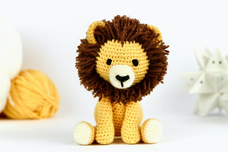 May include: A crocheted lion toy with a brown mane, yellow body, and white face. The lion is sitting with its legs crossed and has black eyes and a black nose.