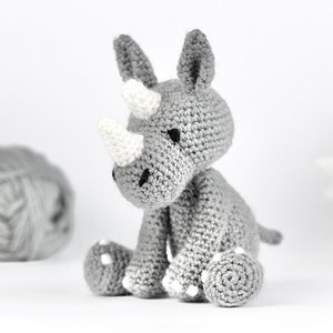 Pode incluir: Um rinoceronte de pel&uacute;cia cinza crochetado com chifres brancos e olhos pretos. O rinoceronte est&aacute; sentado em uma superf&iacute;cie branca.
