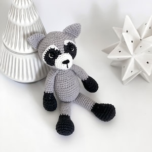 Puede incluir: Un animal de peluche de mapache de punto gris con ojos, nariz y pies negros. El mapache est&aacute; sentado sobre una superficie blanca.