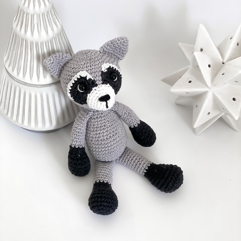Puede incluir: Un animal de peluche de mapache de punto gris con ojos, nariz y pies negros. El mapache est&aacute; sentado sobre una superficie blanca.