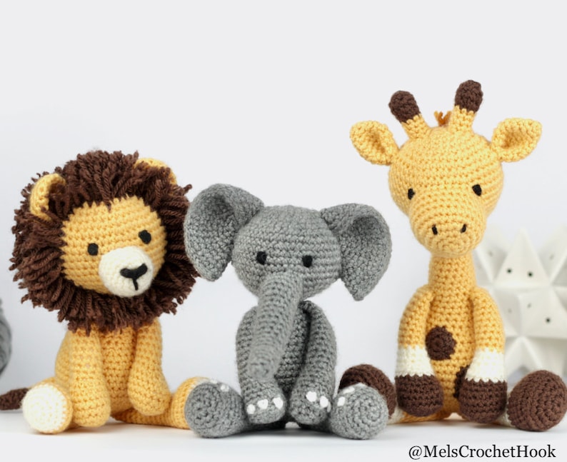 May include: Three crocheted stuffed animals: a yellow giraffe, a gray elephant, and a yellow lion with a brown mane. The animals are sitting on a white surface.