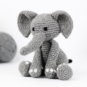 May include: A gray crocheted elephant toy with black eyes and white accents on the feet. The elephant is sitting on a white surface.
