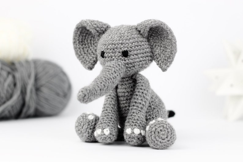 May include: A gray crocheted elephant toy with black eyes and white accents on the feet. The elephant is sitting on a white surface.