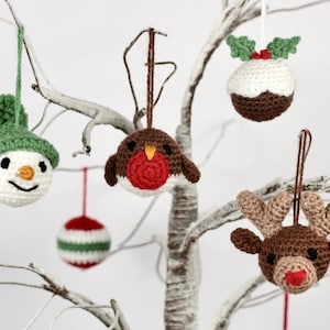 May include: Five crocheted Christmas decorations hanging on a white branch. The decorations are a snowman, a robin, a Christmas pudding, a reindeer, and a striped bauble.