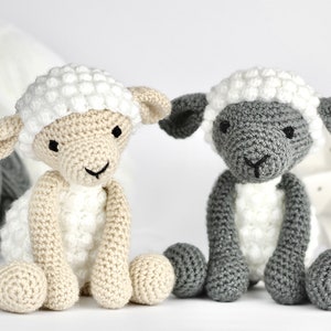 May include: Two crocheted sheep toys, one white and one gray, sitting on a white surface. Both sheep have white faces and black eyes.