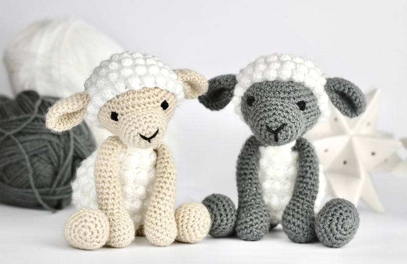 May include: Two crocheted sheep toys, one white and one gray, sitting on a white surface. Both sheep have white faces and black eyes.