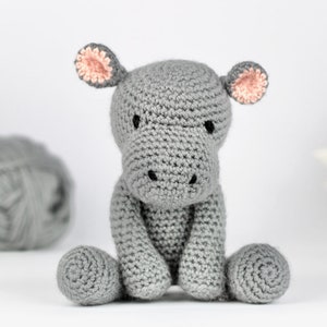 May include: A gray crocheted hippopotamus toy with pink ears. The hippopotamus is sitting on a white surface.