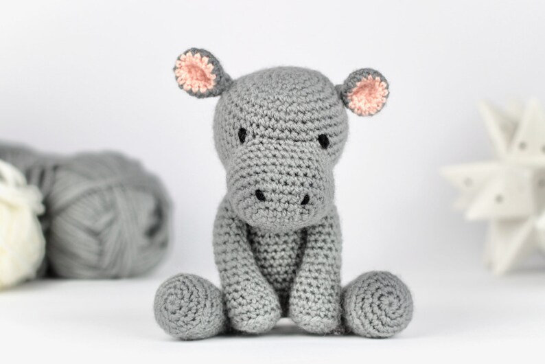 May include: A gray crocheted hippopotamus toy with pink ears. The hippopotamus is sitting on a white surface.