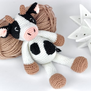 May include: A handmade crochet cow toy featuring a white body with black spots, a pink snout, and light brown hooves and horns. The cow has closed eyes stitched in black thread and small pink-lined ears, crafted with a textured yarn finish.