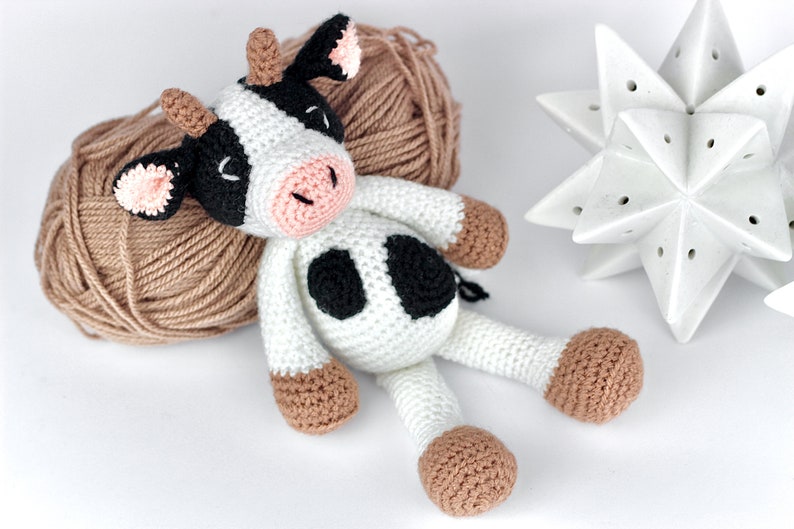 May include: A handmade crochet cow toy featuring a white body with black spots, a pink snout, and light brown hooves and horns. The cow has closed eyes stitched in black thread and small pink-lined ears, crafted with a textured yarn finish.
